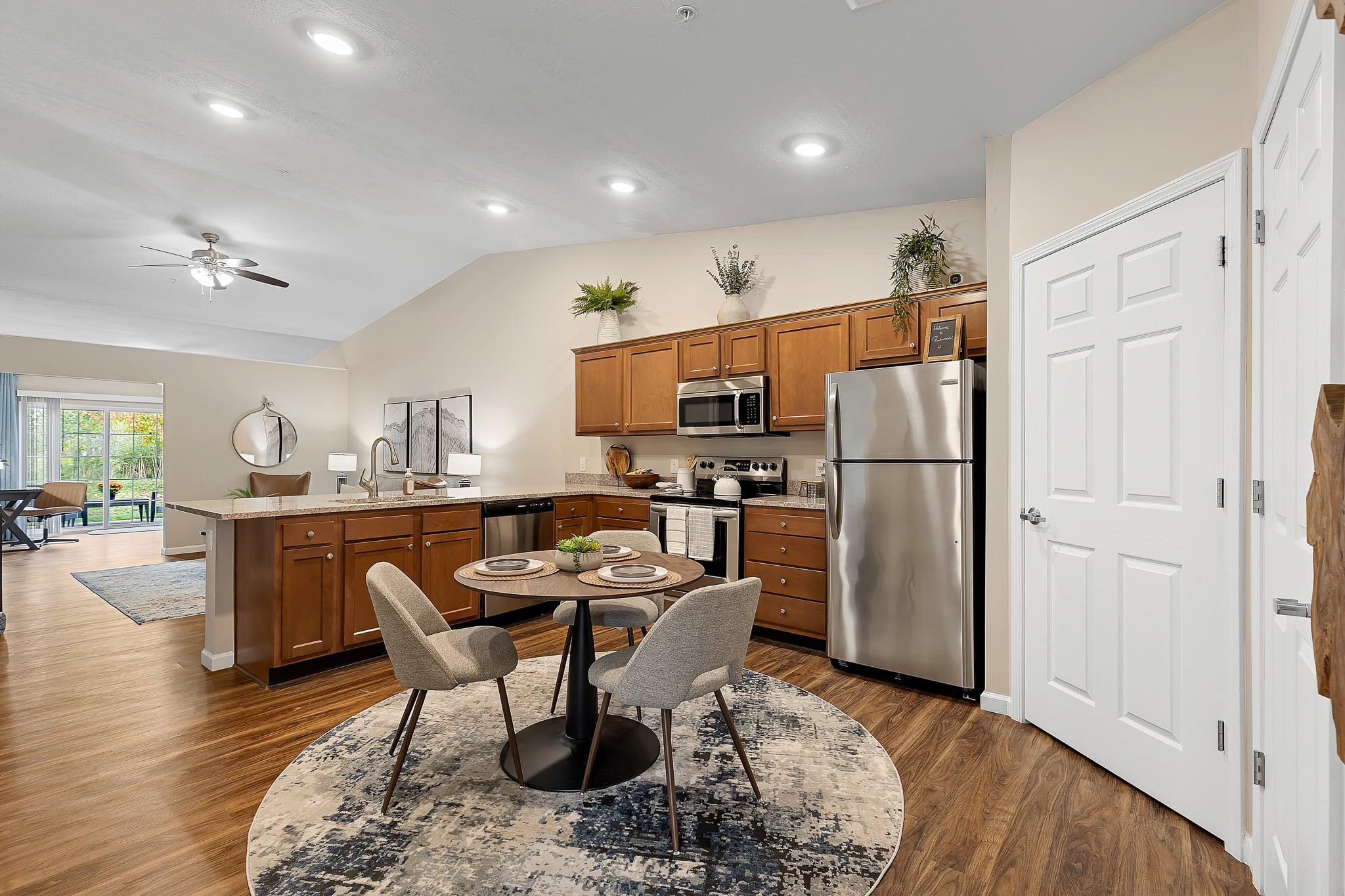 Modern open kitchen with wood cabinets, stainless steel appliances, a round dining table with two chairs, and a rug. Adjacent living and dining areas are visible in the background.