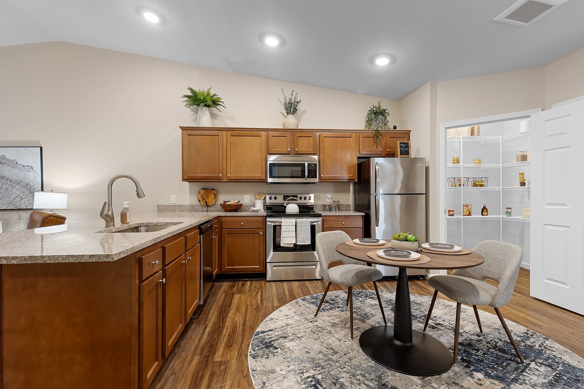 Modern kitchen with wooden cabinets, stainless steel appliances, a round dining table with three chairs, and an open pantry stocked with food items.