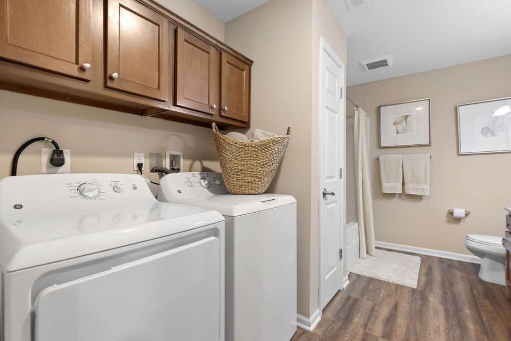 Laundry room with washer and dryer, wicker basket on top, wooden cabinets above, and an adjacent bathroom with towels and framed art on the wall.