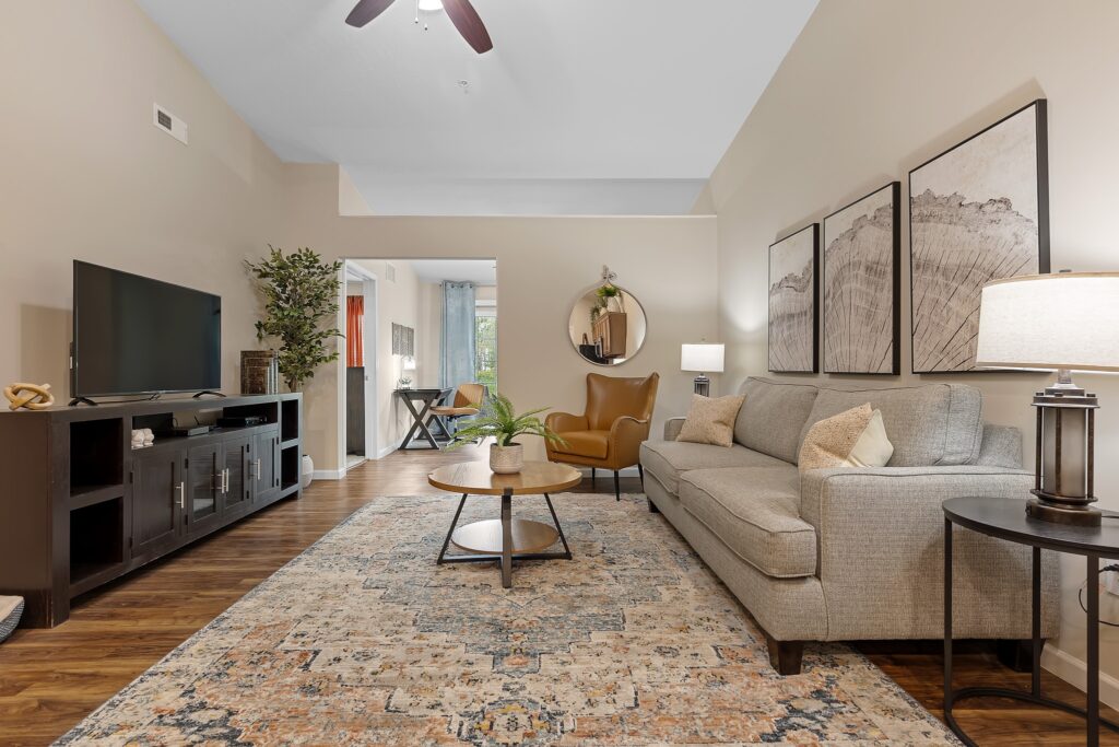 A modern living room with a gray sofa, brown armchair, round coffee table, TV on a console, wall art, and a patterned rug on wooden flooring. A small desk is visible in the next room.