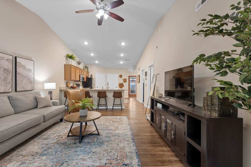 Open-plan living room and kitchen with a sofa, coffee table, TV console, bar stools, indoor plants, wall art, and wooden flooring under recessed and ceiling fan lighting.