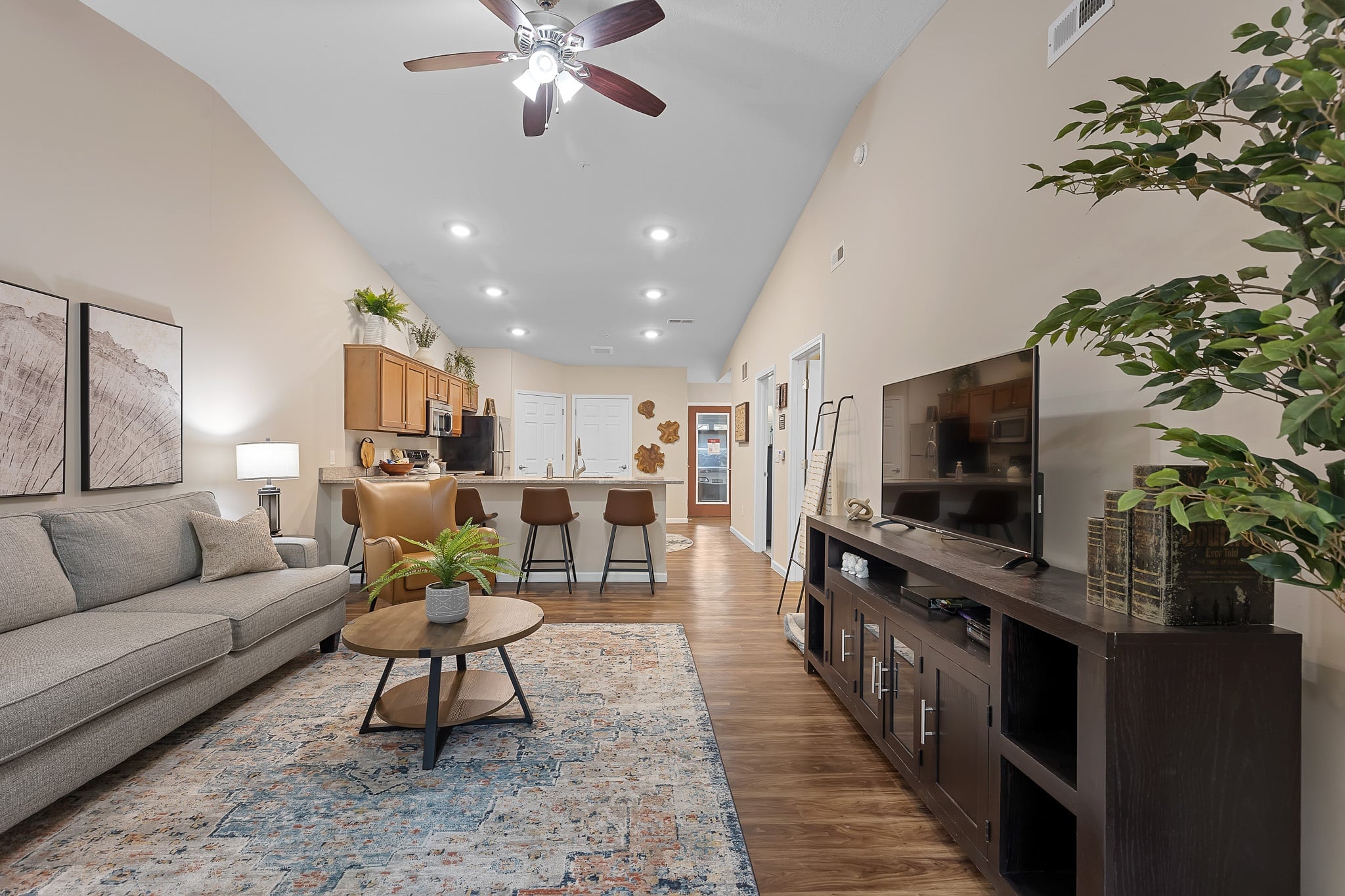 Open-plan living room and kitchen with a sofa, coffee table, TV console, bar stools, indoor plants, wall art, and wooden flooring under recessed and ceiling fan lighting.