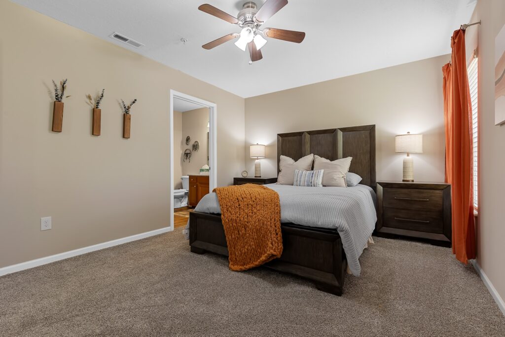 Bedroom with a double bed, gray bedding, orange throw, two nightstands with lamps, beige walls, wall decor, carpeted floor, and an open door to a bathroom.