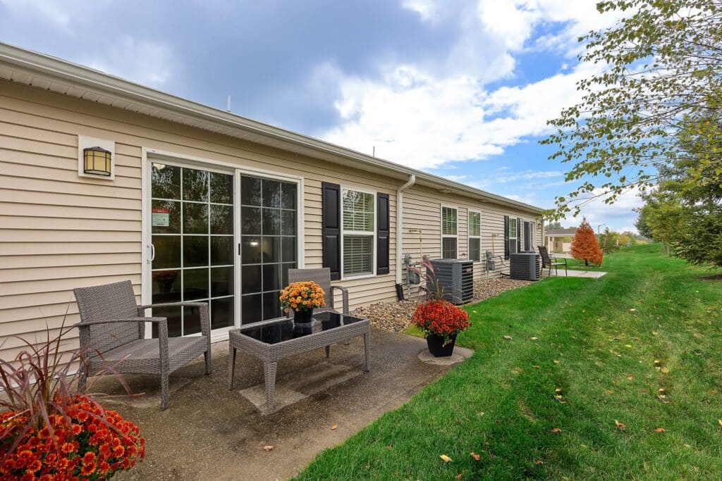 A row of beige single-story homes with sliding glass doors, small patios, outdoor chairs, potted plants, and a grassy lawn under a partly cloudy sky.