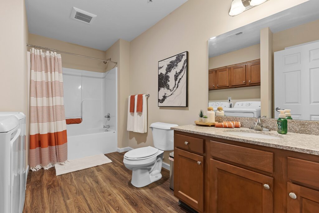 Modern bathroom with bathtub, toilet, wood cabinets, and a large mirror; stacked washer and dryer visible in the adjoining laundry area. Orange and white towels and decor.
