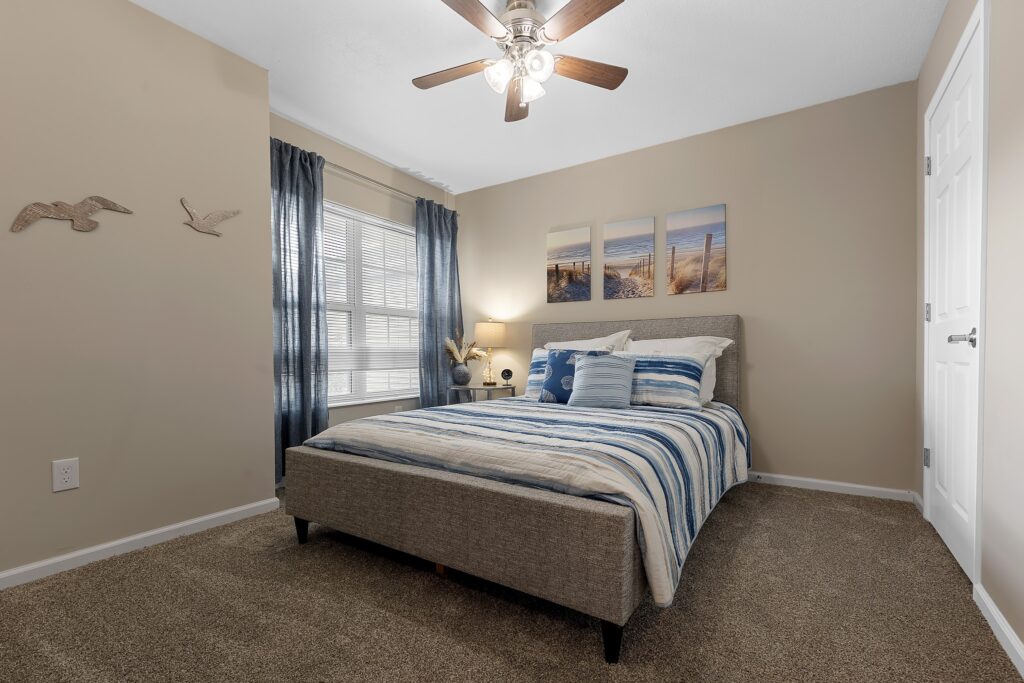 A neatly made bed with striped bedding is centered in a carpeted bedroom, featuring wall art, blue curtains, a window, and a ceiling fan with lights.