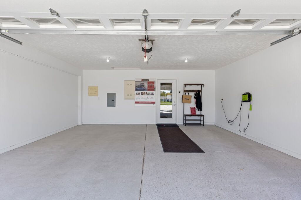 Open view of a clean, empty two-car garage with a door to the house, wall-mounted organizer, coat rack, and electric panel on the left wall.