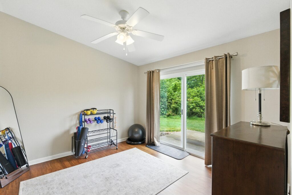 A home gym room with exercise equipment, a floor mat, a dresser, a full-length mirror, ceiling fan, and sliding glass doors opening to a green backyard.