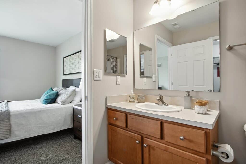 A bathroom with a single sink vanity, wooden cabinets, and wall mirrors is shown, with a view into an adjacent bedroom featuring a neatly made bed with pillows.