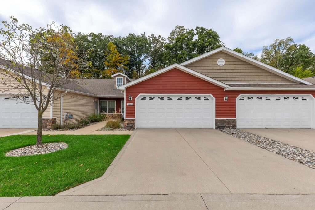 A duplex with tan and red siding, attached double garages, concrete driveways, and a small front lawn with a tree, set against a backdrop of trees.
