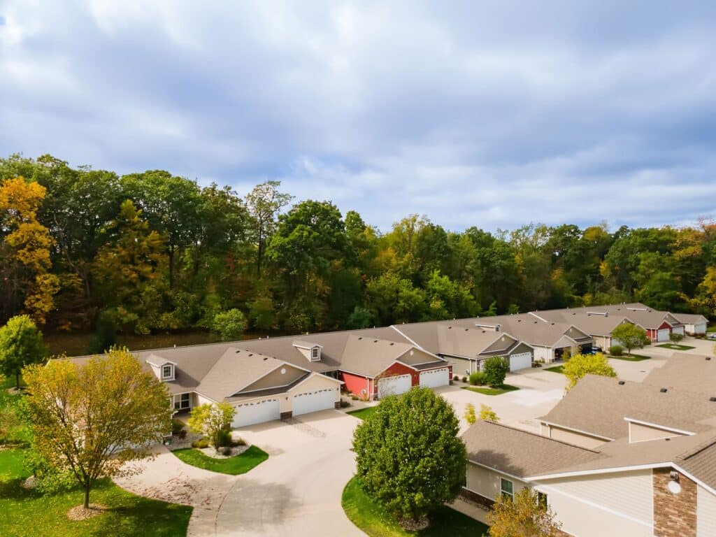 A row of suburban houses with attached garages lines a quiet street, bordered by trees with early autumn foliage under a partly cloudy sky.