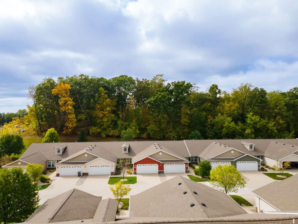 Row of townhouses with garages and driveways, trees with autumn foliage in the background, and cloudy sky overhead.
