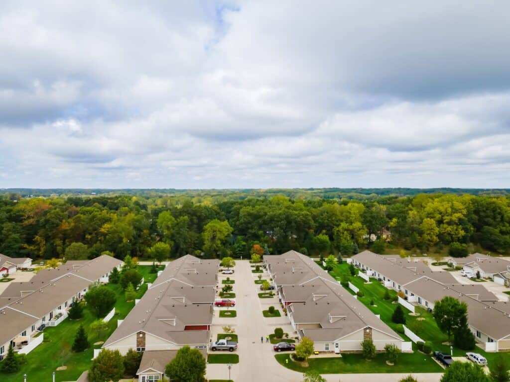 Aerial view of a suburban neighborhood with rows of houses, green lawns, trees, and a forested area in the background under a cloudy sky.