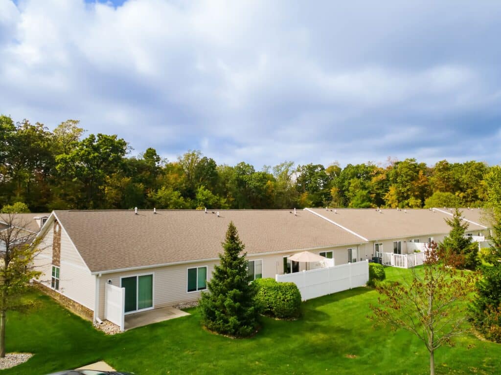 Row of single-story beige townhouses with fenced patios, set in a grassy area with trees in the background under a cloudy sky.