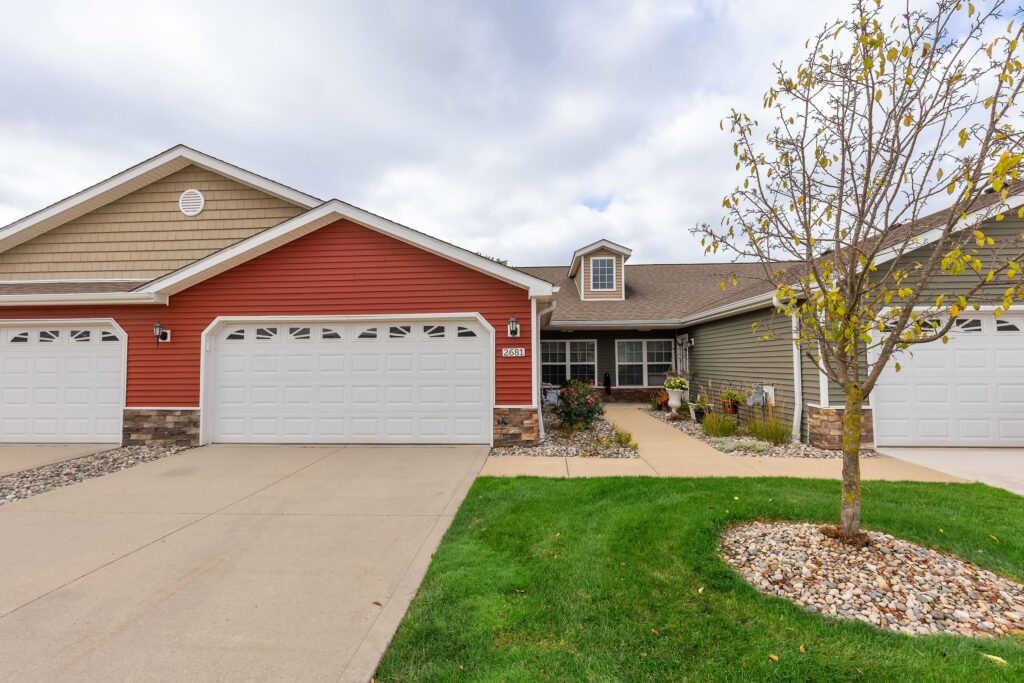 A duplex with two white garage doors, red and beige siding, stone accents, a small front yard, and a young tree on a cloudy day.