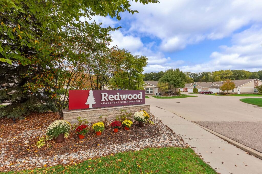 A sign reading "Redwood Apartment Neighborhoods" stands by a landscaped area with flowers near a residential street lined with houses and trees.
