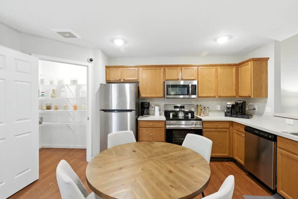 Modern kitchen with stainless steel appliances, wooden cabinets, round dining table with four chairs, and a visible pantry with organized shelves in the background.
