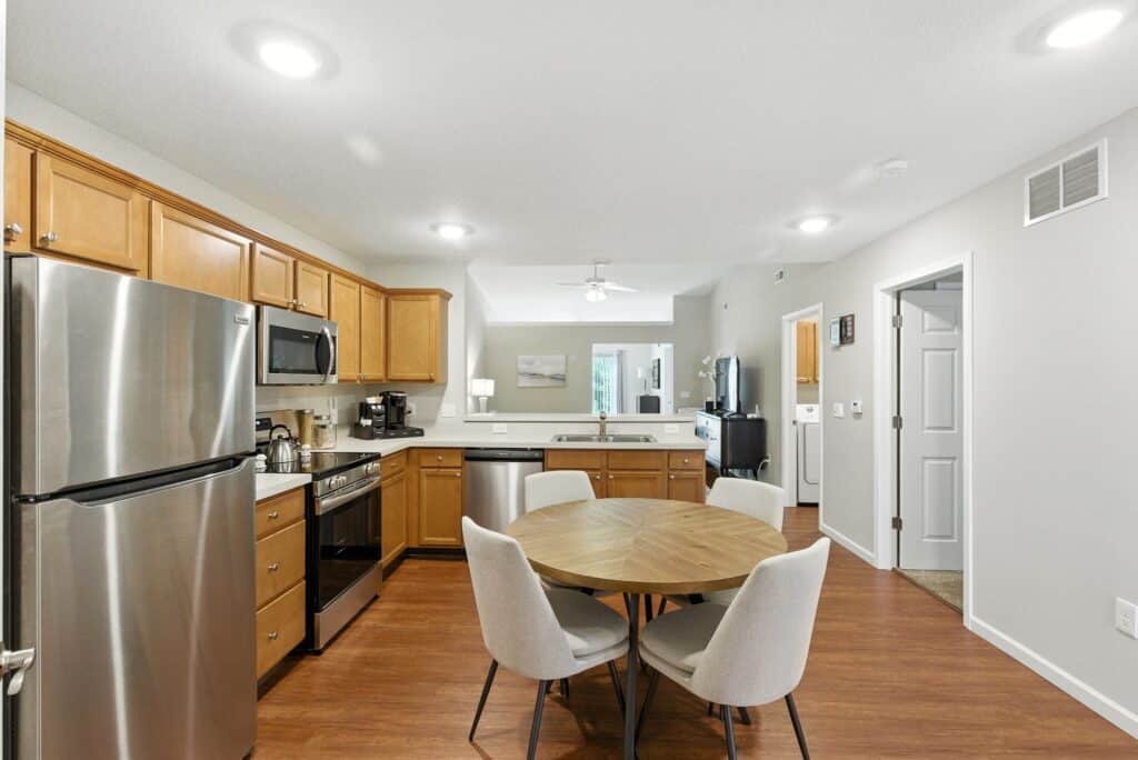 Modern kitchen with light wood cabinets, stainless steel appliances, a round dining table with four chairs, and hardwood flooring. Adjacent to an open living area and hallway.