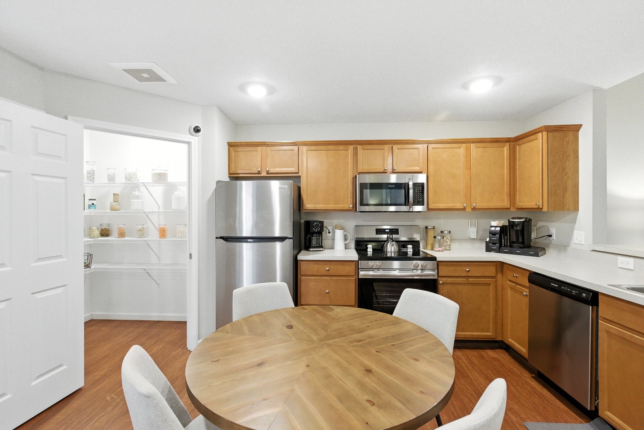 Modern kitchen with stainless steel appliances, wooden cabinets, round dining table with four chairs, and a visible pantry with organized shelves in the background.