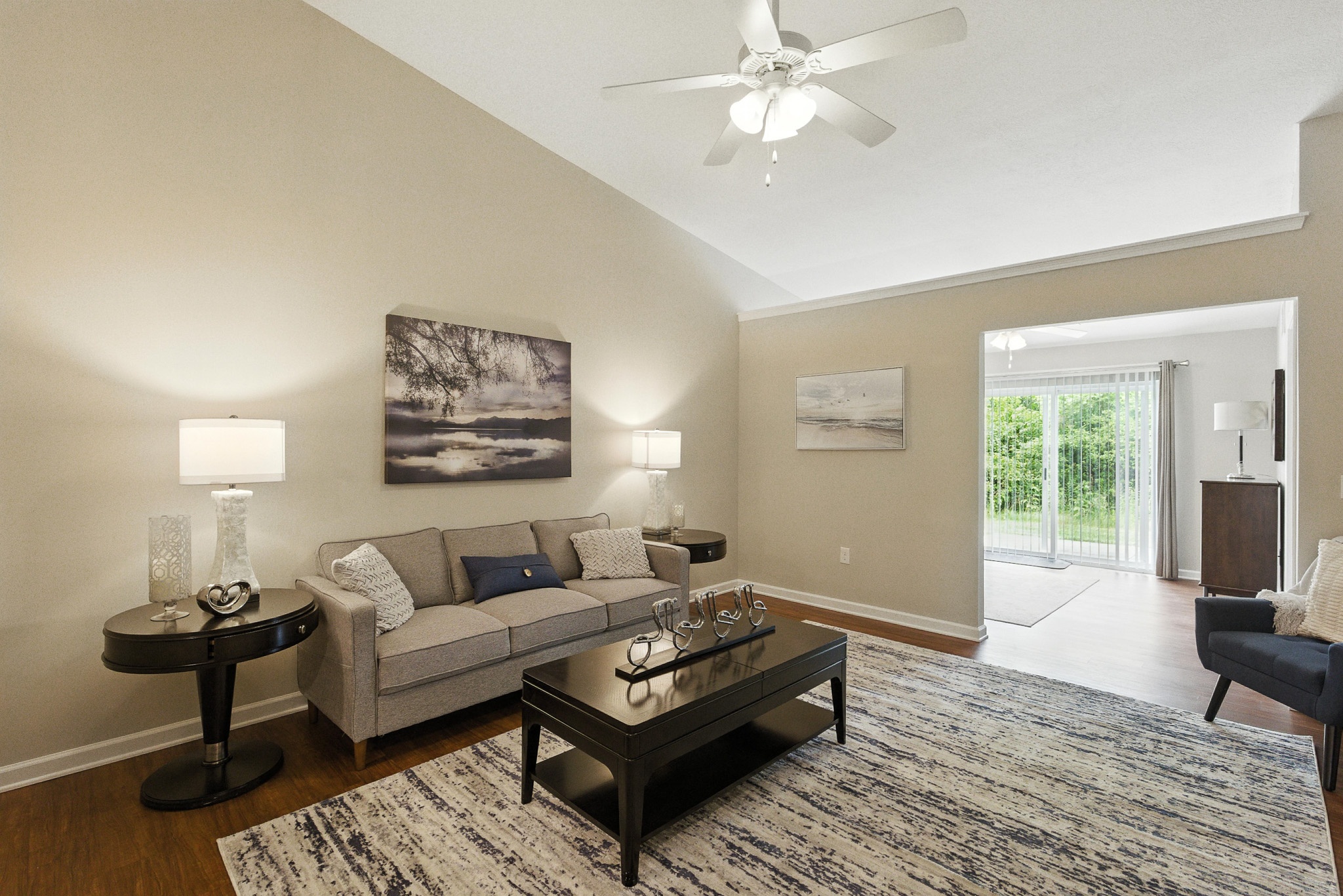 A modern living room with a gray sofa, coffee table, area rug, side tables with lamps, wall art, and large windowed doors opening to a patio with greenery outside.