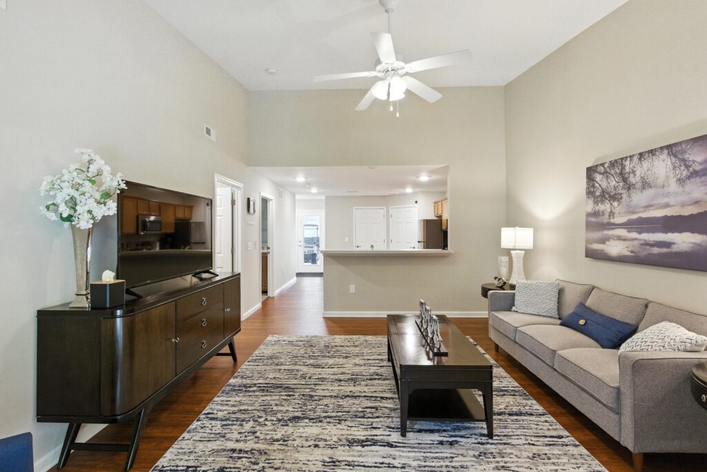 Living room with a gray sofa, coffee table, TV on console, patterned rug, wall art, ceiling fan, and view into a bright kitchen area.