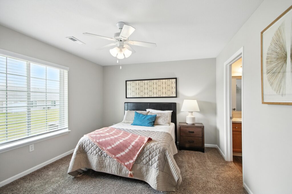 A bedroom with a bed, a side table with a lamp, a large window with blinds, and an open door leading to a bathroom. The walls are light gray and the room has carpeted flooring.
