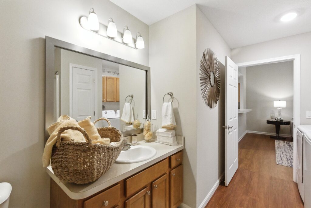 A bathroom with a single sink, wooden vanity, large mirror, basket of towels, stacked toiletries, and towel rings; a hallway with wood flooring is visible through the open door.