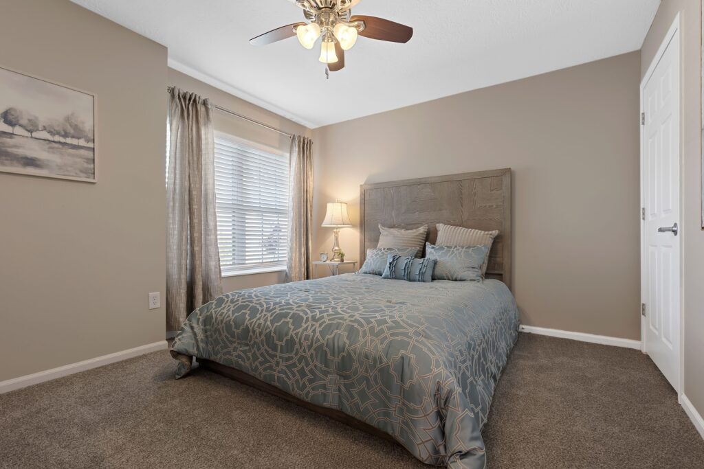A neatly made bed with patterned blue bedding in a carpeted bedroom, featuring a window with curtains, a nightstand with a lamp, and a ceiling fan with lights.