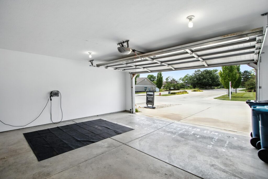 Clean, empty garage interior with a partially open door, a wall-mounted device with a cord, a black mat on the floor, and a view of a suburban street outside.
