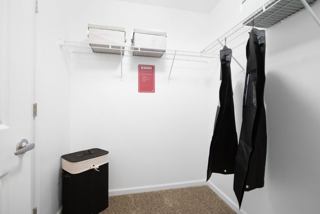 A clean, empty walk-in closet with wire shelves, two hanging black garment bags, storage bins, a laundry hamper, and a sign on the back wall.