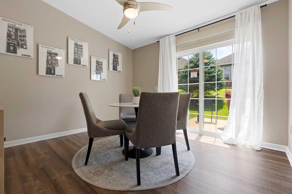 A dining area with a round table and four chairs sits on a rug near large windows with sheer curtains, overlooking a green yard.
