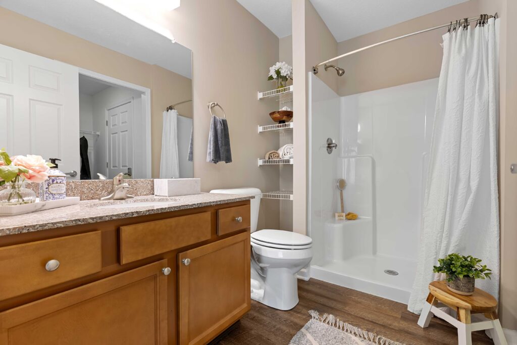 Modern bathroom with a wood vanity, granite countertop, toilet, walk-in shower with white curtain, wall shelves, and decorative plants on a wood floor.