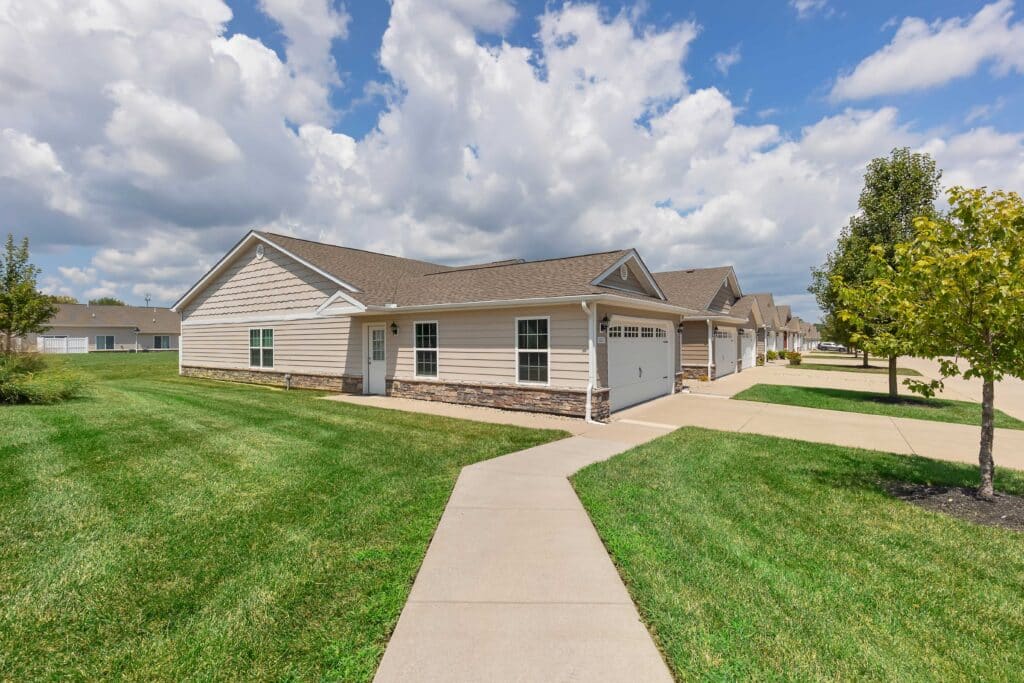Single-story houses with attached garages line a residential street, bordered by green lawns, sidewalks, and young trees under a partly cloudy sky.