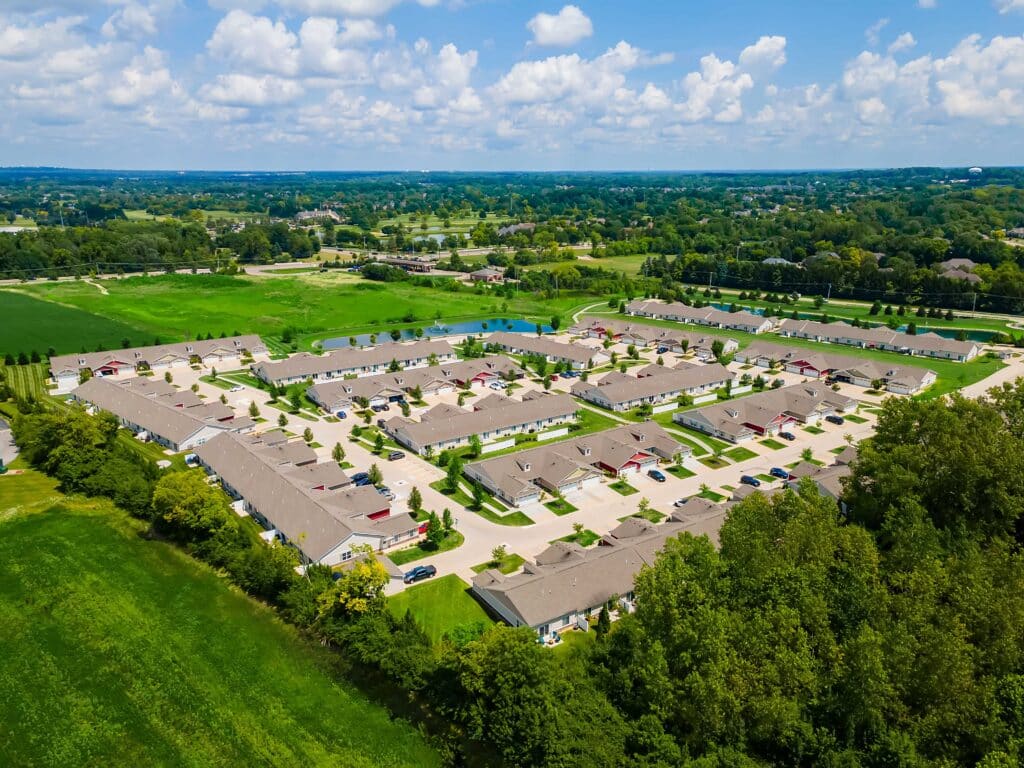 Aerial view of a suburban residential neighborhood with rows of houses, green lawns, trees, and surrounding open fields under a partly cloudy sky.