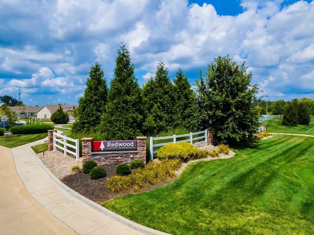 Landscaped entrance to the Redwood residential community featuring a sign, white fence, green lawn, trees, and cloudy blue sky.