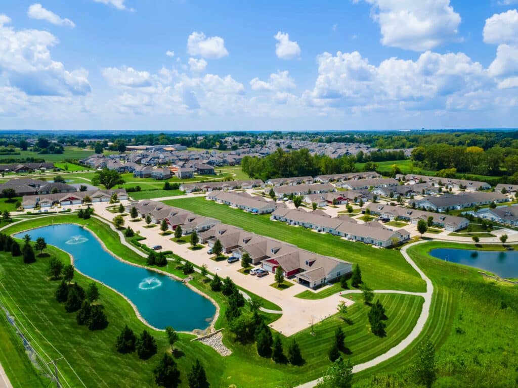 Aerial view of a suburban neighborhood with rows of houses, green lawns, two ponds with fountains, and surrounding trees under a partly cloudy sky.