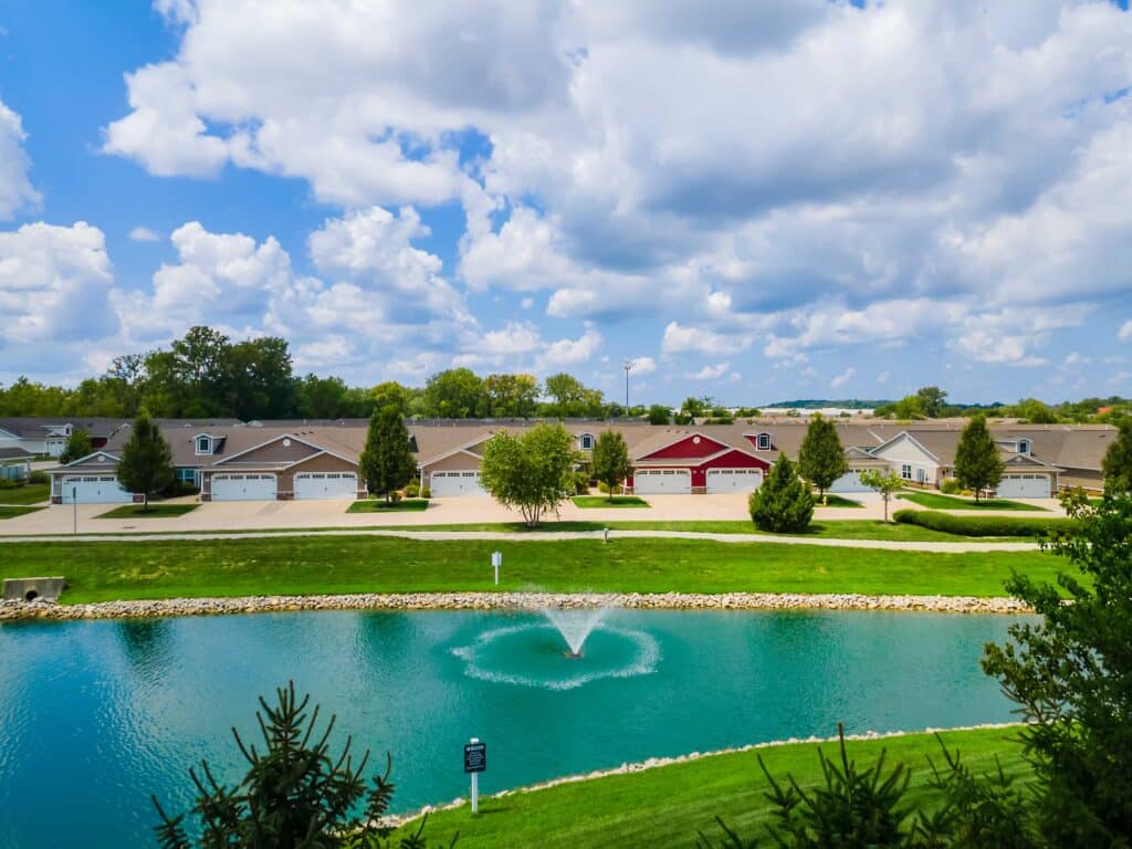 A row of single-story houses with garages faces a small pond with a fountain, under a partly cloudy sky.