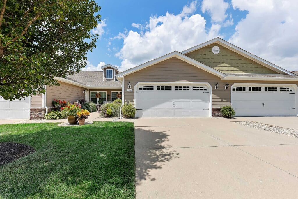 Single-story suburban house with a three-car garage, beige siding, manicured front lawn, potted plants, and a tree on the left under a partly cloudy sky.