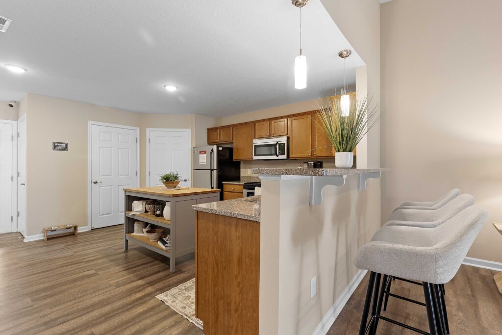A modern kitchen with wood cabinets, stainless steel appliances, an island, bar seating with two stools, and light brown walls.