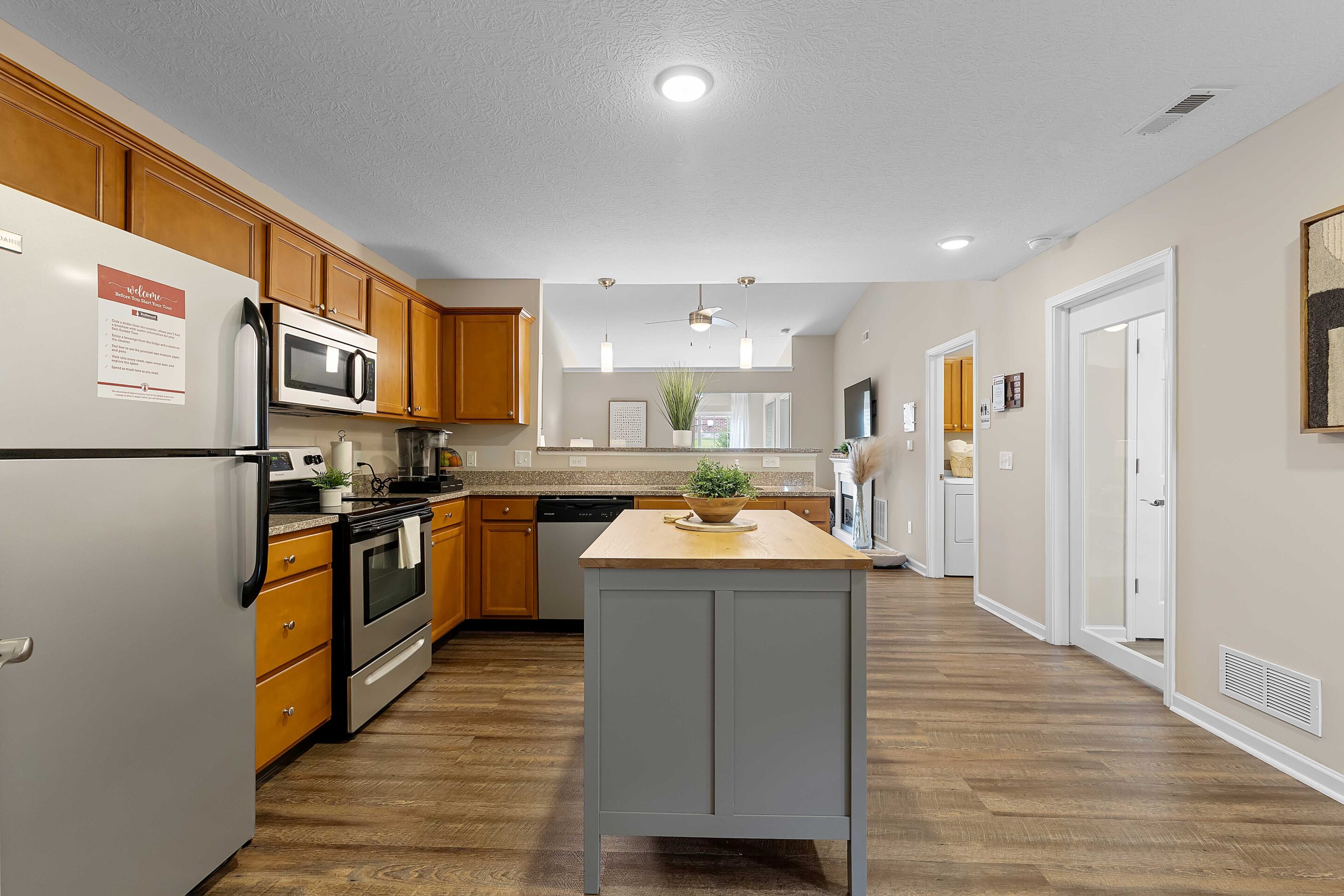 Modern kitchen with stainless steel appliances, wooden cabinets, central island, and wood flooring. A living area and hallway are visible in the background.