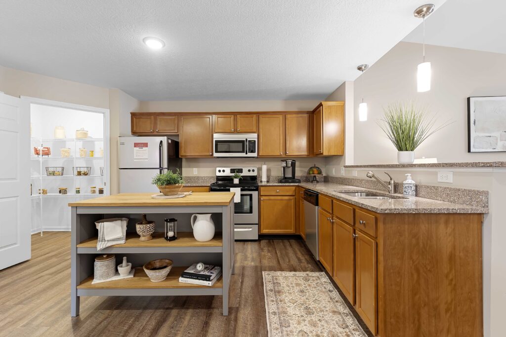 Modern kitchen with wooden cabinets, stainless steel appliances, a center island with shelves, and a view of a pantry with open door. Countertops have a plant, sink, and decorative items.