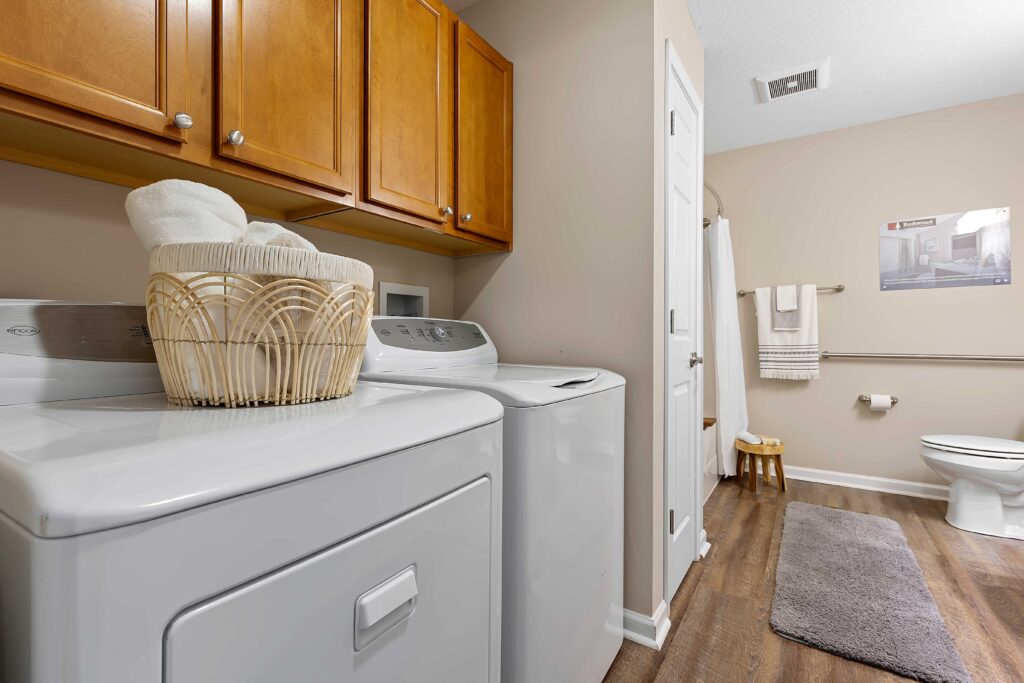 Laundry room with a washer and dryer, basket of towels on top, wooden cabinets above, bathroom area with toilet, towel rack, and gray rug on the floor.
