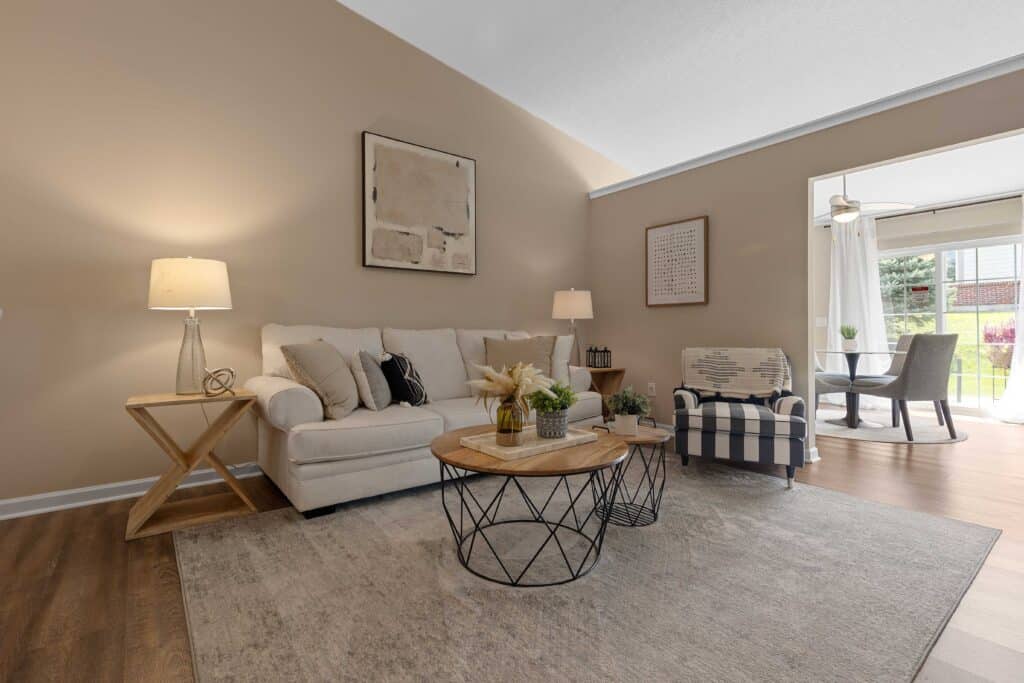 A neutral-toned living room with a beige sofa, striped armchair, round coffee table, lamps, and wall art; dining area visible in the background near glass doors.