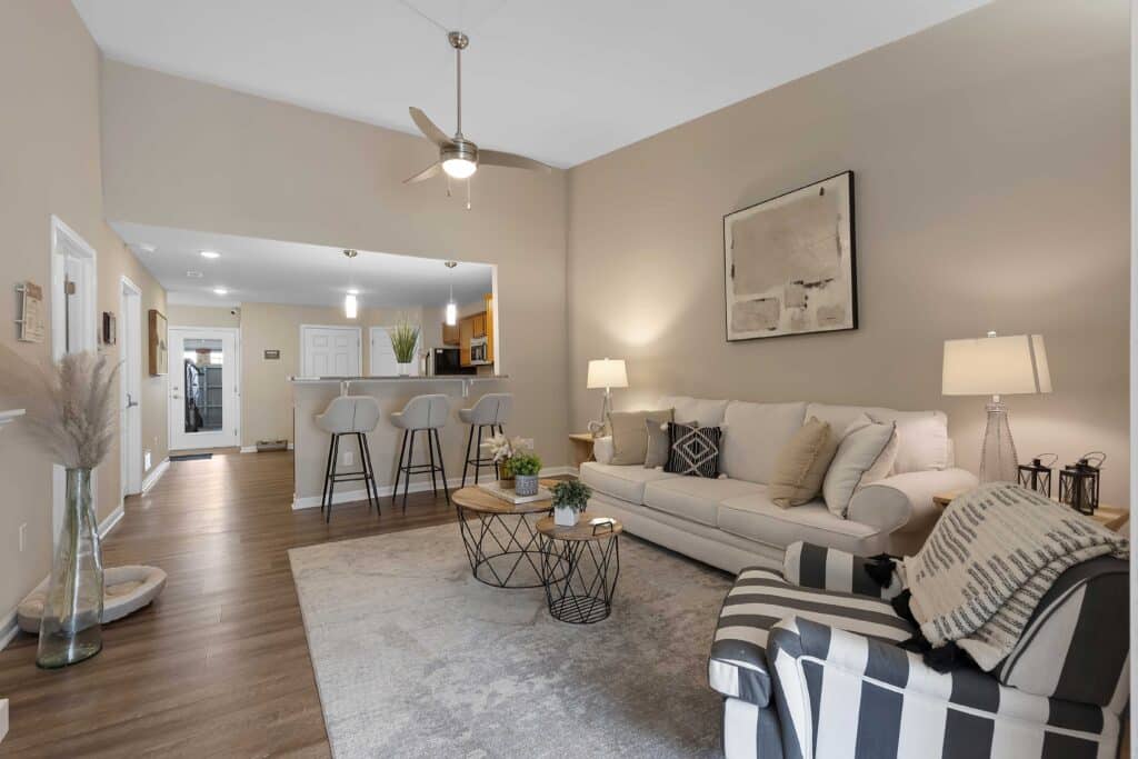 Modern living room with a neutral palette, featuring a beige sofa, striped armchair, round coffee tables, barstools at a kitchen counter, and minimalist wall art.