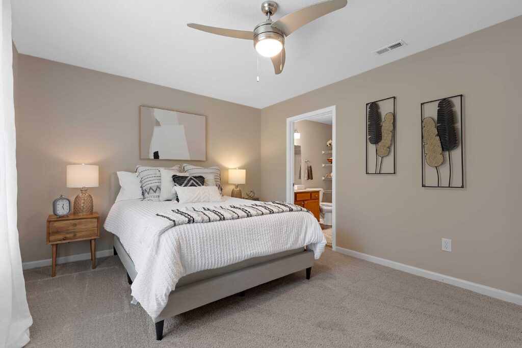 A neatly made bed with white bedding is centered in a beige bedroom. Nightstand with lamps, abstract wall art, and an open doorway to a bathroom are visible.