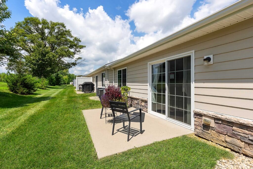 Small patio with two chairs and a table outside a beige single-story house, next to a lawn and sliding glass doors, under a partly cloudy sky.