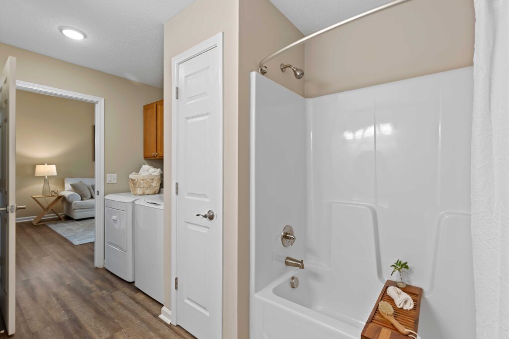 Bathroom with a white bathtub and shower, a wooden tray, and a view into a laundry area with a washer, dryer, and shelves, and a living room in the background.
