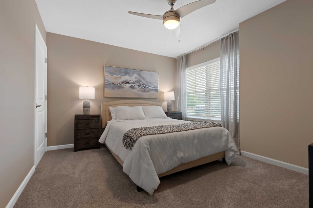 A neatly made bed with two nightstands, lamps, and a landscape painting in a beige-walled bedroom with carpeted floor and a ceiling fan.
