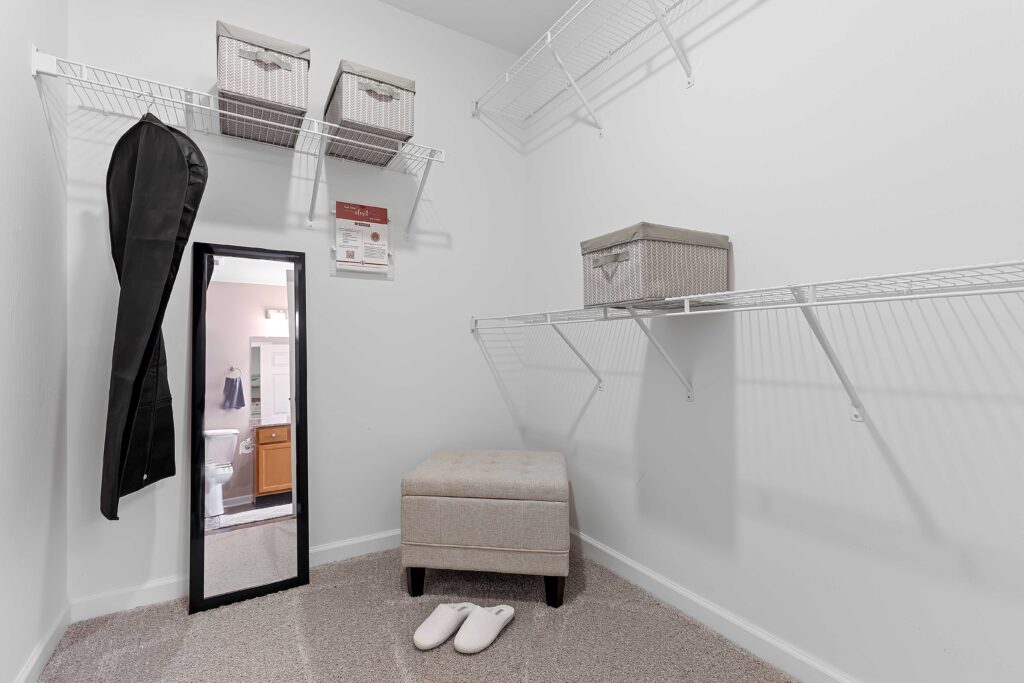 Walk-in closet with wire shelves, a mirror, a storage ottoman, three storage bins, a clothing bag, and a pair of white slippers on carpeted floor. Bathroom visible through open door.