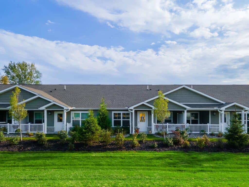 Single-story residential building with a covered porch, several windows, small trees, shrubs, and a well-maintained lawn under a partly cloudy sky.